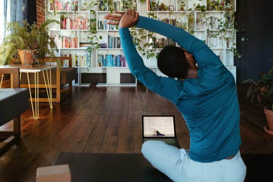 Man gör yogaklass hemma med dator och yogaklass online. Mannen har på sig mjuka kläder, en blå långärmad tröja och ljusa byxor. I bakgrunden är en vit bokhylla på en hel vägg med böcker och växter. Det står även möbler i rummet. Mannen sitter på en mörk yogamatta och bredvid ligger en yogakloss av kork.