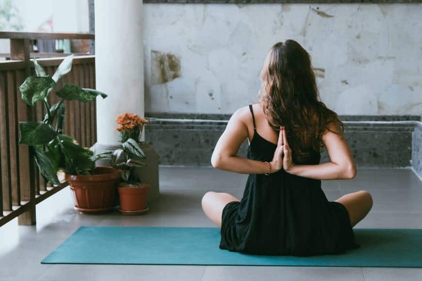 Ung kvinna sitter med en svart klänning på en blå yogamatta på en ballkong. Hon sitter ner med benen i kors och håller sina händer knäppta bakom ryggen. Hennes mörkbruna hår är utsläppt och bilden är tagen bakifrån. På ballkongen står också tre krukor med gröna växter och en med blommor. Yoga på yogamatta utomhus