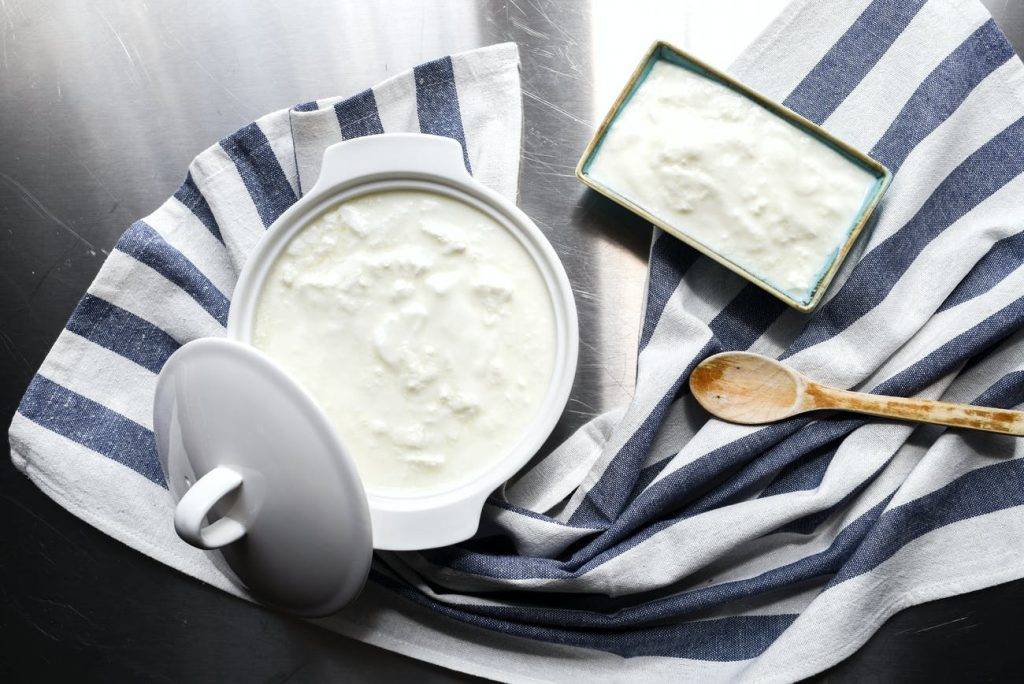 Hemmagjord yoghurt i gryta och en skål med yoghurt bredvid. Grytan är av keramik och locket ligger på sidan. Grytan är full med krämig, hemmagjord yoghurt. Bredvid finns en fyrkantig skål fylld med yoghurt och en träsked bredvid. Grytan och skålen sitter på en randing kökshandduk. Skapar mejeriprodukter inflammation