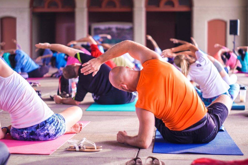 Full yogaklass. Bilden är tagen bakifrån yogaklass där många deltagare sitter nära varandra på yoga mattor. Utövarna av yoga strechar sidan av kroppen sittandes ner. Framtill syns högtalare och en yogalärare som sitter på en liten scen.