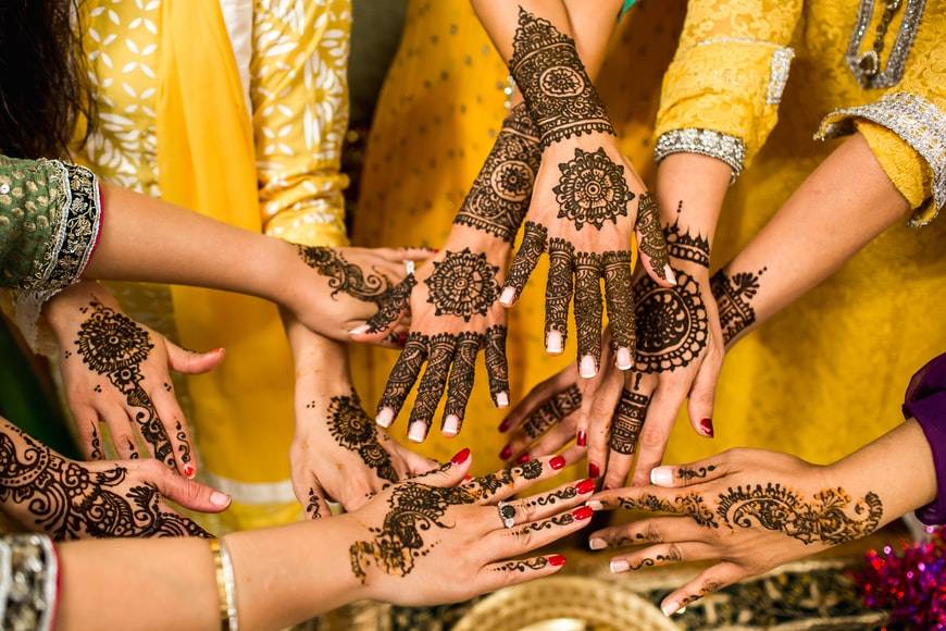 Flera kvinnor håller fram sina armar och händer med hennatatueringar. Kvinnorna har gula saris och en kvinna har en grön sari. Bilden är tagen så att enbart kvinnornas armar syns, med en del av deras överkropp och saris. Kvinnorna har händerna och armarna tatuerade med tradionella indiska mönster av henna. Samt indiska smycken.