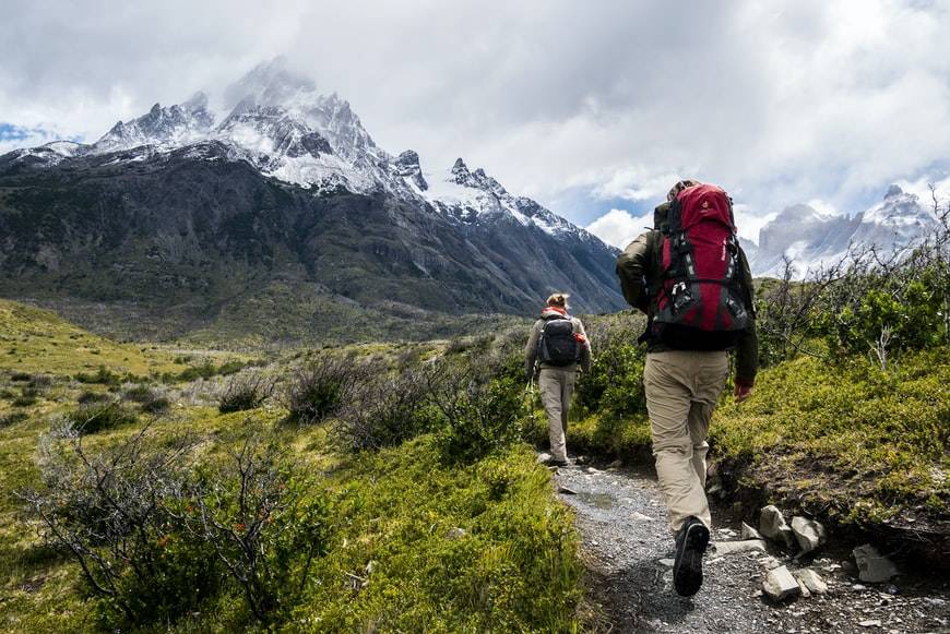 Två personer är på hike och vandrar i berg. De har ryggsäckar på ryggarna och vandringsskor. På bergstopparna ligger ett tunt lager med snö och naturen är lite grön. 