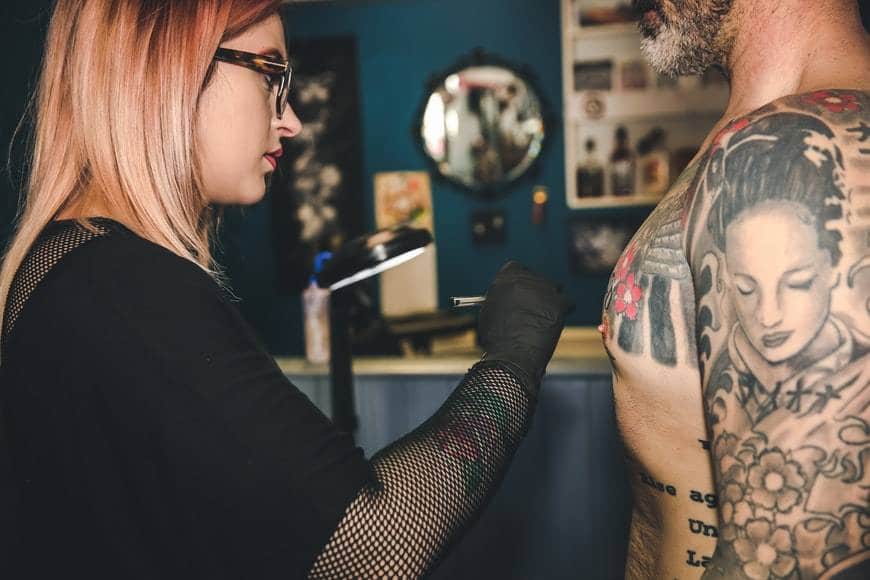 Kvinnlig tatuerare målar innan tatueringen börjar på mannen som ska tatueras. Mannen har redan många tatueringar på överkroppen. Kvinnan har en penna i handen och svarta plasthandskar. Målar med penna innan tatueringsfärger