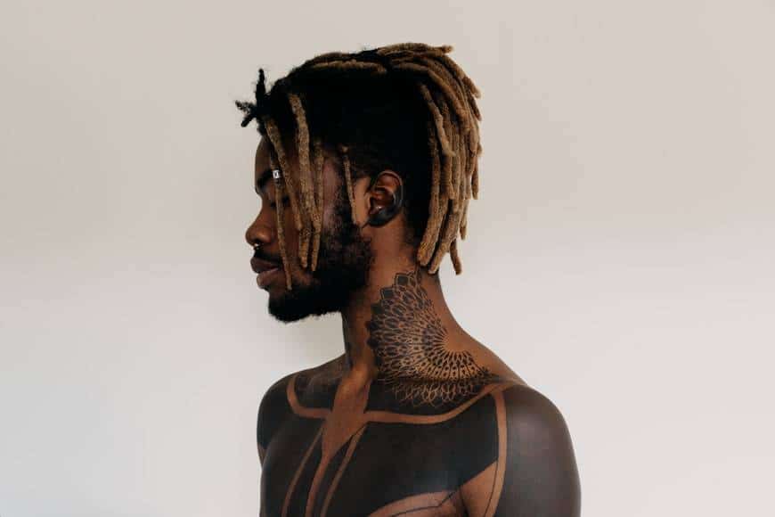 Man med tatuerad överkropp. Bilden är tagen på mannens överkropp där han har svarta tatueringar över hela bröstet, armarna och upp på halsen. Mannen har dreads i page och även tatuering på örat som syns på bilden. Svarta tatueringsfärger