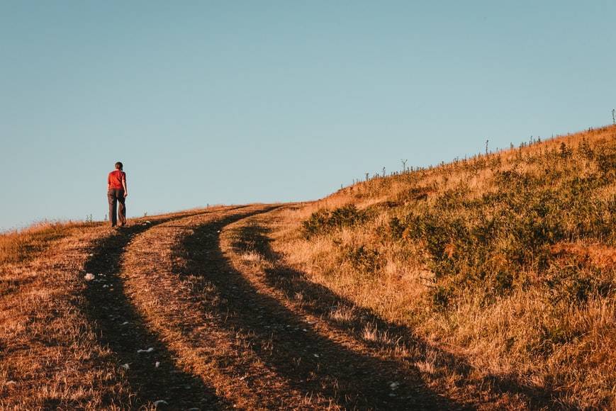 Promenad i uppförsbacka utomhus. En person går uppför i en backe med brunt, torrt gräs och klarblå himmel. Det ser ut att vara tidigt på morgonen eller sent på eftermiddagen.