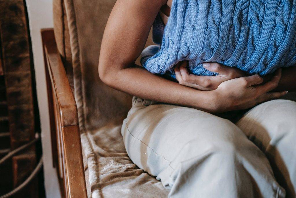 Kvinna håller för magen som att den gör ont. Bilden är tagen på kvinnan från axlar neråt. Hon sitter på en stol och har en blå kortärmad tröja på sig som visar hennes armar, vilka hon har omsvept runt sin mage och lätt framåtlutat överkropp. Ansamling vätksa och magknip vid PMS