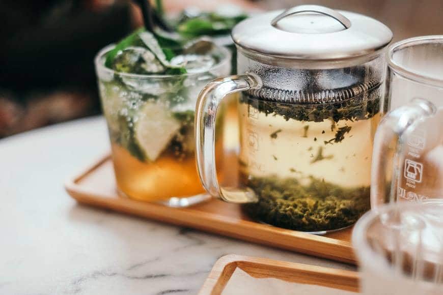 Varmt och kallt örtte. Det står två glas med kallt örtte med gröna örter, lime och is på en bricka tillsammans med en liten tekanna i glas som har örter i sig. Bredvid står en tom liten glaskanna på brickan. Brickan står på ett bord