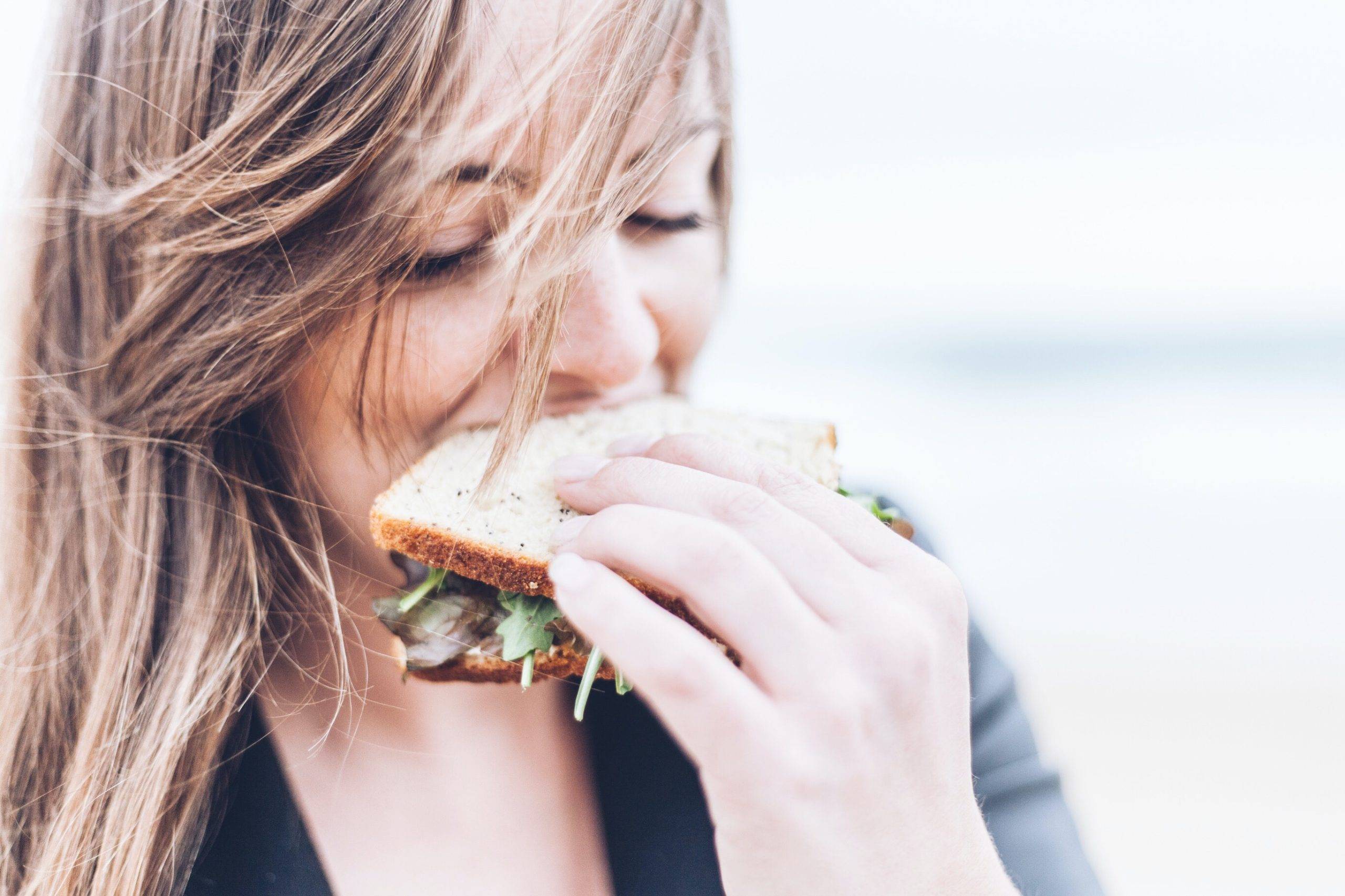 Kvinna äter en dubbelmacka. Bilden är tagen på kvinnans ansikte när hon håller en smörgås i handen och tar ett bett. Hon har brunt utsläppt hår och äter utomhus
