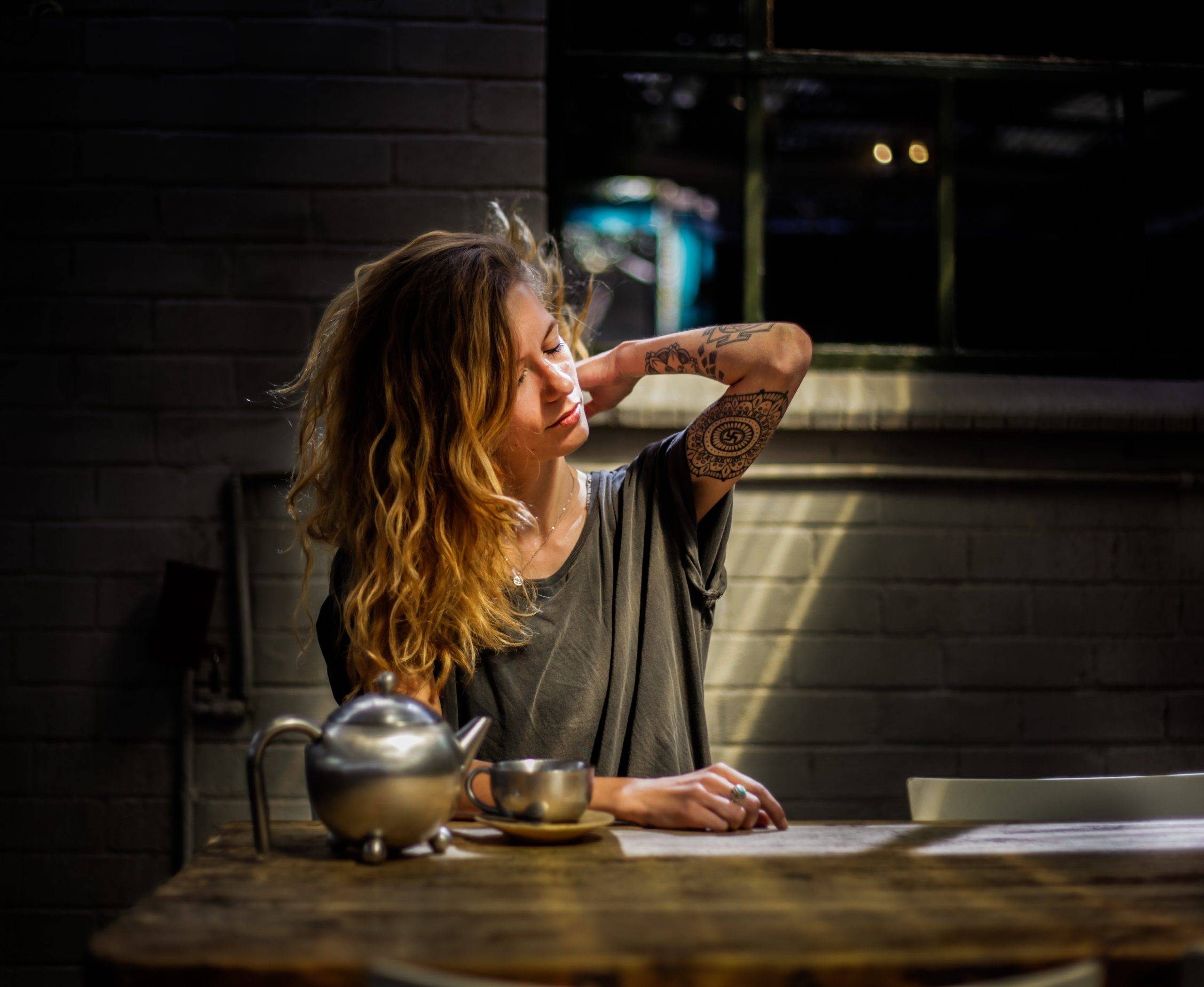 Kvinna sitter vid köksbord i mörkret på kvällen med en tekanna och kopp framför sig på bordet. Kvinnan har sin ena arm uppsträckt som har tatueringar på sig.
