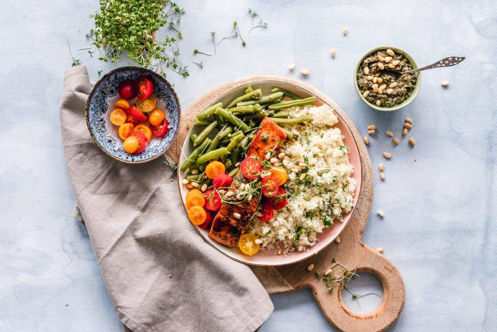 Tallrik med vit quinoa, stekt lax, gröna bönor ocg körsbärstomater. Tallriken står på en träskärbärda och bredvid ligger en linnehandduk, en söt skål med körsbärstomater i halvor och en liten skål med pesto med pinjenötter ovanpå. Livsmedel rika på melatonin