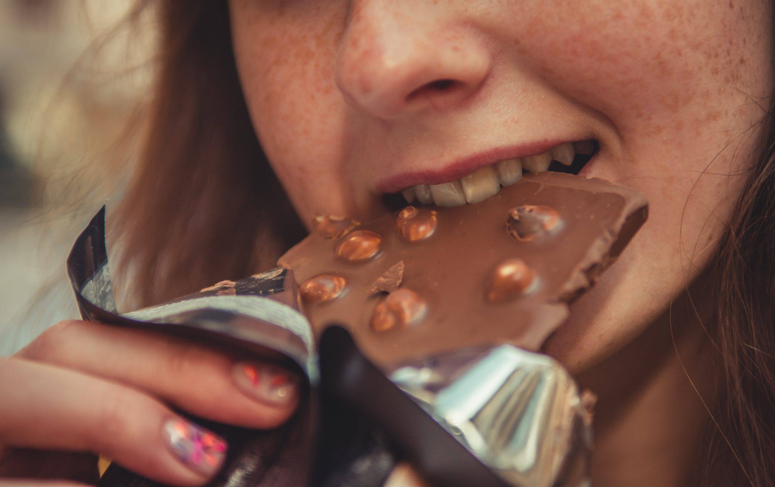 Närbild på kvinna med fräknar som äter en chokladkaka, mjölkchoklad med hela hasselnötter. Pappret är neddraget från chokladkakan och hon tar ett stort bett i mjölkchokladen.