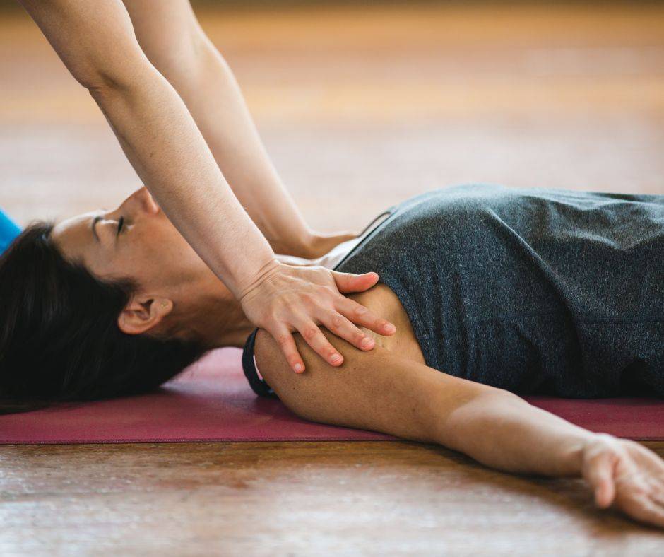 Kvinna ligger ner på en yogamatta med armarna ut på sidorna och slutna ögon. En annan kvinnan håller sina händer och på kvinnans axlar och trycker ner dem i golvet. Breathwork