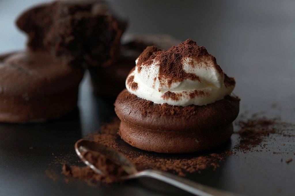 Rund chokladkaka med grädde ovanpå och kakaopulver stött ovanpå. I bakgrunden står tre chokladkakor till och framför kakan ligger en sked.