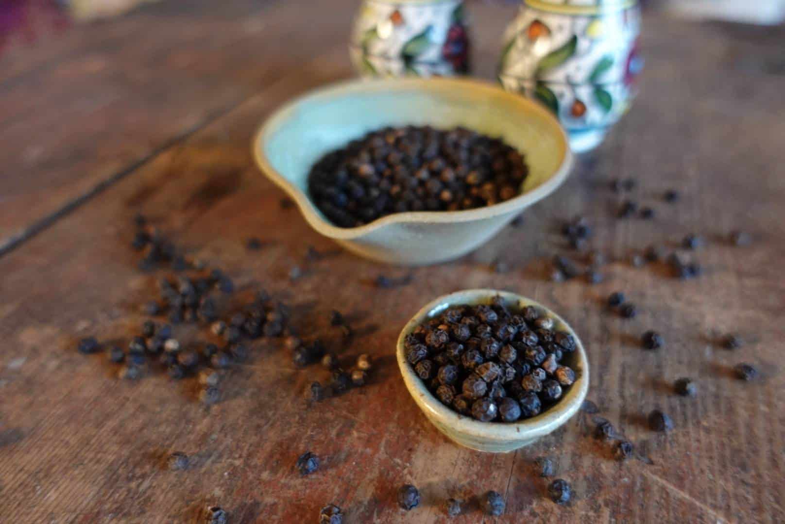 Svartpeppar i små keramikskålar som står på ett träbord. Det ligger hela korn av svartpeppar utspritt runt skålarna och i bakgrunden syns två salt och pepparkvar i keramik.