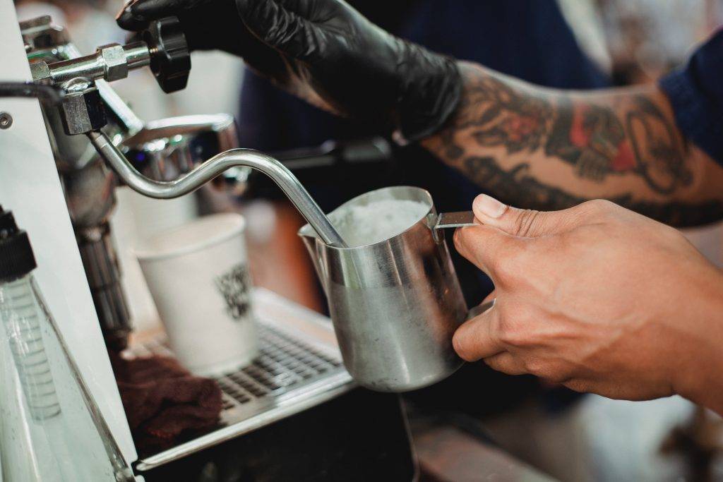 Barista gör kaffe på cafe och skummar mjölk i espressomaskinen. Han har en svart plasthandske på ena handen med tatueringar på underarmen. Det står en pappersmugg på espressomaskinen.