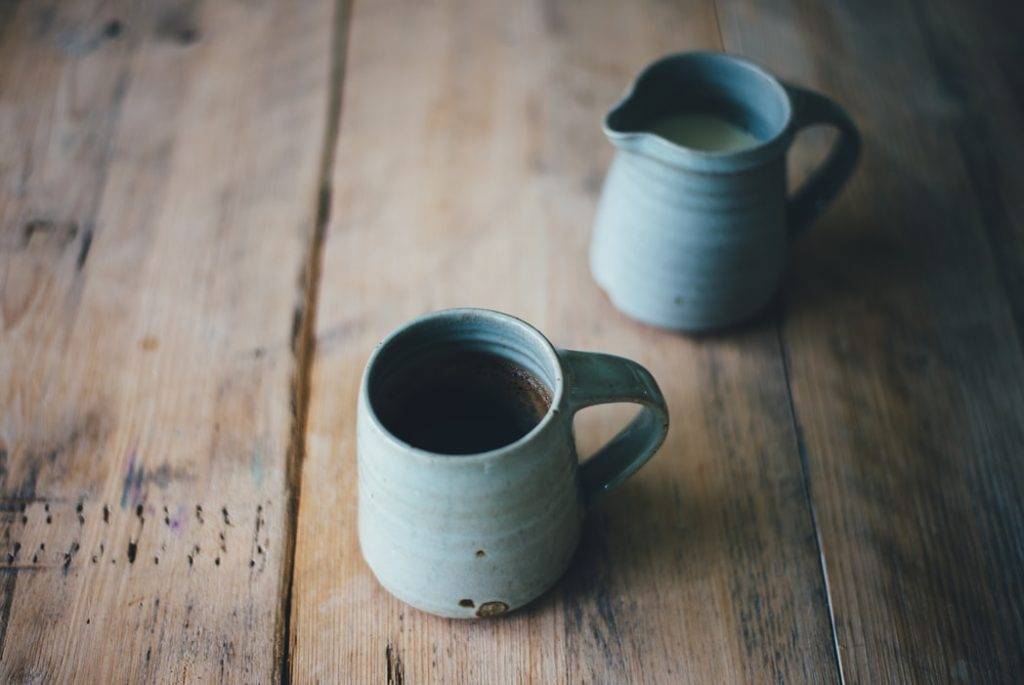 Blå keramikkopp med svart kaffe och blå mjölkkanna i keramik med mjölk står på ett rustikt träbord. Bilden är tagen i närbild på keramiken