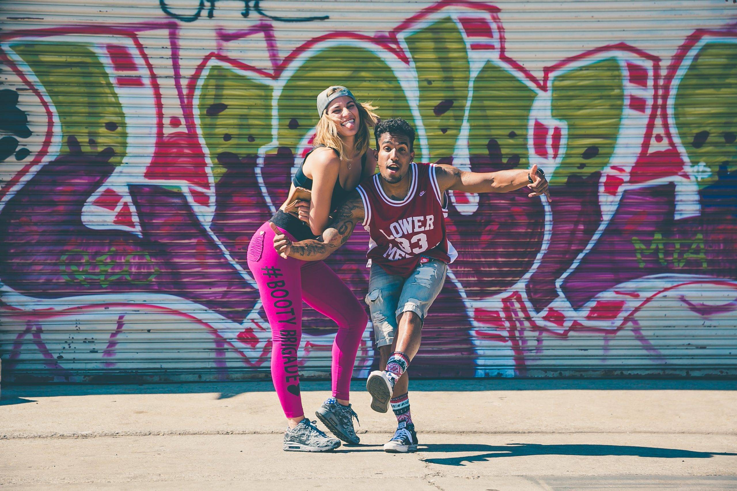 Ung kvinna och man står framför en graffitivägg utomhus med färgglada kläder och i danssteg. Båda tittar in mot kameran med glad uppsyn. Kvinnan har rosa tights. sporttopp och kejp. Mannen har jeansshorts och baskettröja