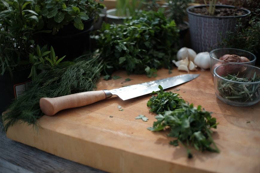 Färska gröna örter i krukor och på skärbräda av trä. På skärbrädan finns också en kniv med träskaft och två glas; ett med charlottenlök och ett med hackade örter. Det ligger vitlöksklyftor vid sidan om.