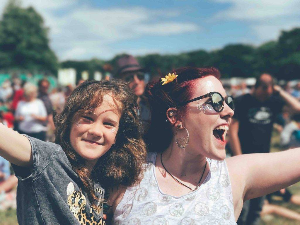 Mamma och dotter skrattar och håller ut armarna. Bilden är tagen i närbild på när mamman håller sin ca 10-åriga dotter i famnen på festival. I bakgrunden syns många människor och vädret är soligt. Mamman ropar glatt och sträcker ut armen medan dottern ler in i kameran med armen utsträckt.