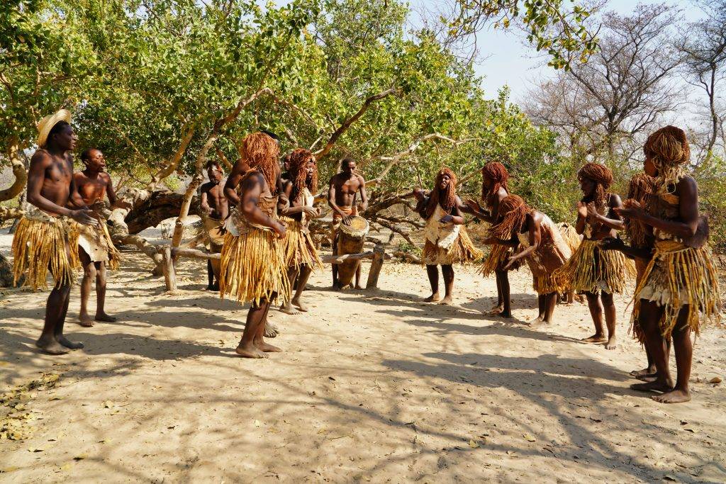 Afrikansk folkstam dansar på stampad lermark. I bakgrunden står gröna träd och personerna står i ring med kjolar och lätta kläder. En man i mitten spelar trumma och de andra klappar händerna och sjunger. Dans för hälsan