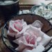 Rosa nyplockade rosor ligger i en skål av keramik på en flätad låg korg på ett träbord. Bredvid är en skål med andra vita blommor och en kopp med te på rosor. Det ligger rosenblad utspritt runt skålarna.