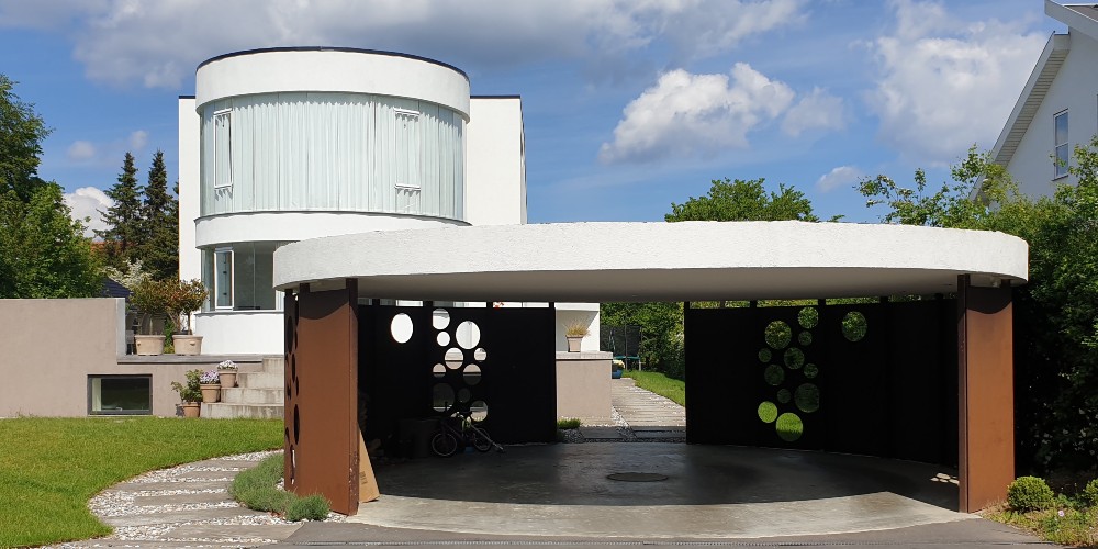 Rund carport i forgrunden som matcher moderhuset, som er arkitekttegnet lavenergihus_designhus i Holte_Beton_metal_staal. Bozel - når typehuse ikke er nok