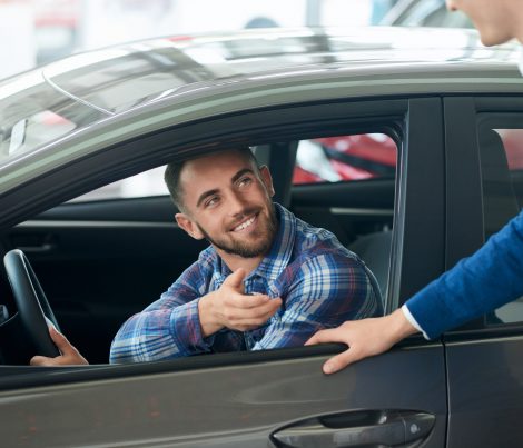 Happy smiling handsome man in shirt sitting in car after successful vehicle buying. Unrecognizable male car seller and young male customer handshake after new silver car purchase.