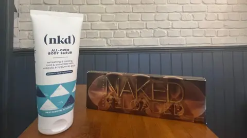 A white tube which says (nkd) all over body scrub on a shelf next to an eye shadow pallet which says Naked Reloaded.