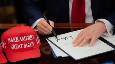 Getty Images US President Donald Trump signs an executive order in the Oval Office at the White House in Washington, DC on 23 April 2025. A red baseball hat embroidered with "Make America Great Again" in white thread sits to the left of the document, which shows Trump's distinctive signature