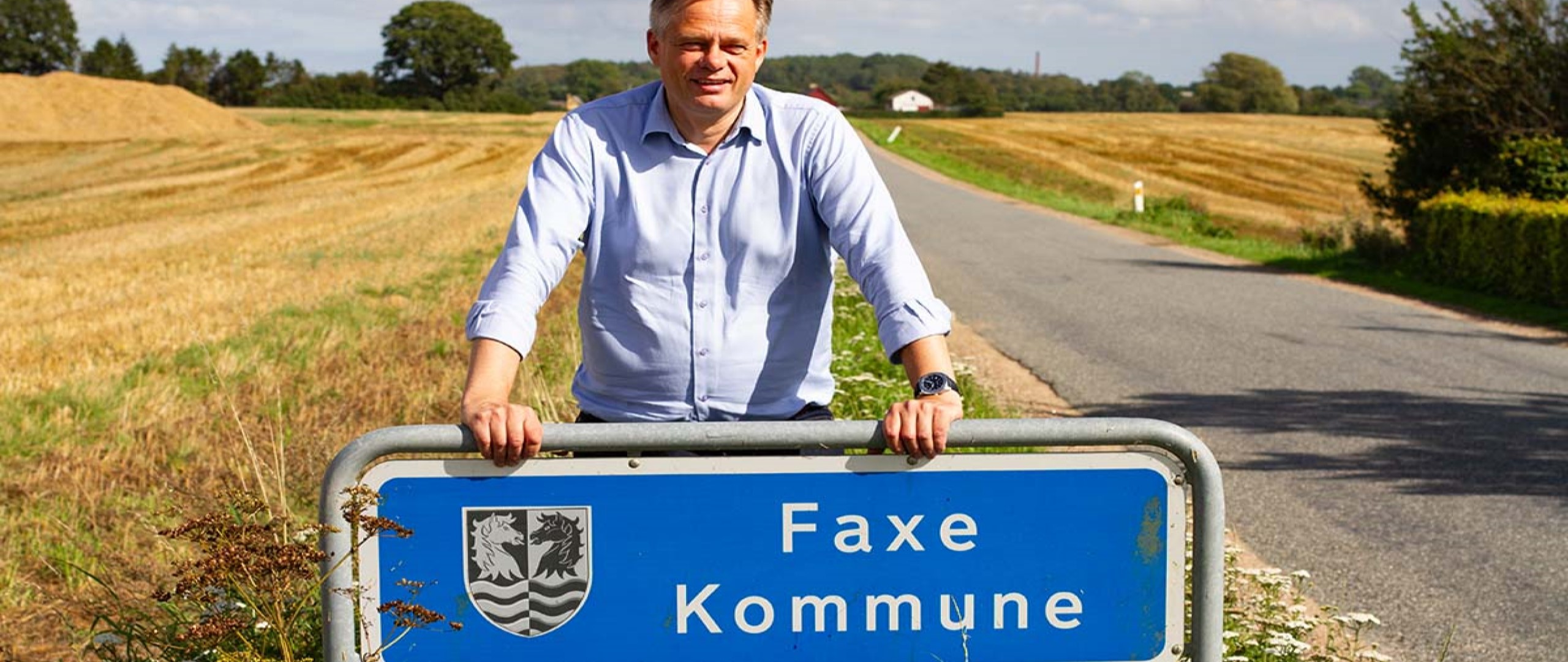 Erhvervskonsulent Peter Fjerring stående, smilende ved et Faxe Kommune byskilt