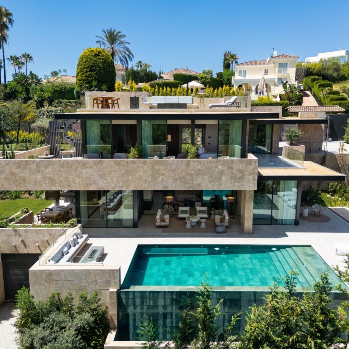 A multi-levelled stone villa with cantilevered levels and terraces. At the front is a rectangular pool and there are other houses, gardens and palm trees in the background