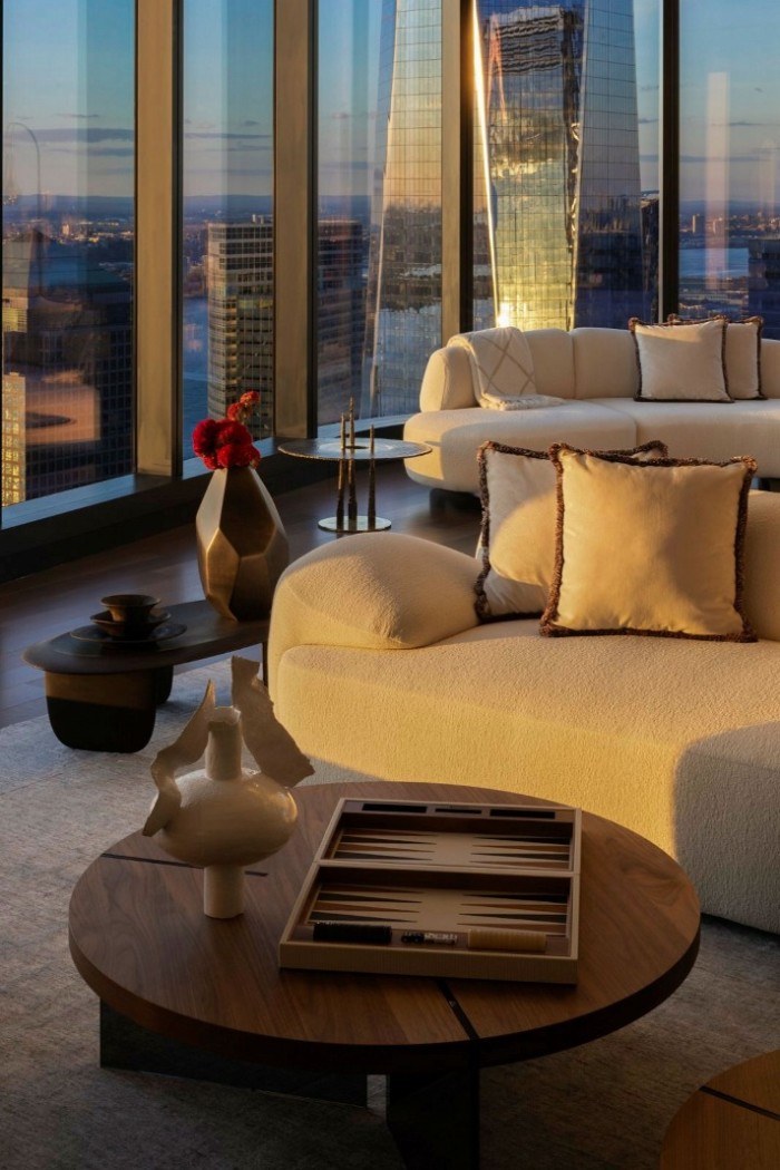 A high-rise living space with curved sofas, a round wooden table, decorative vases, a backgammon set, floor-to-ceiling windows, and a cityscape reflecting on glass buildings