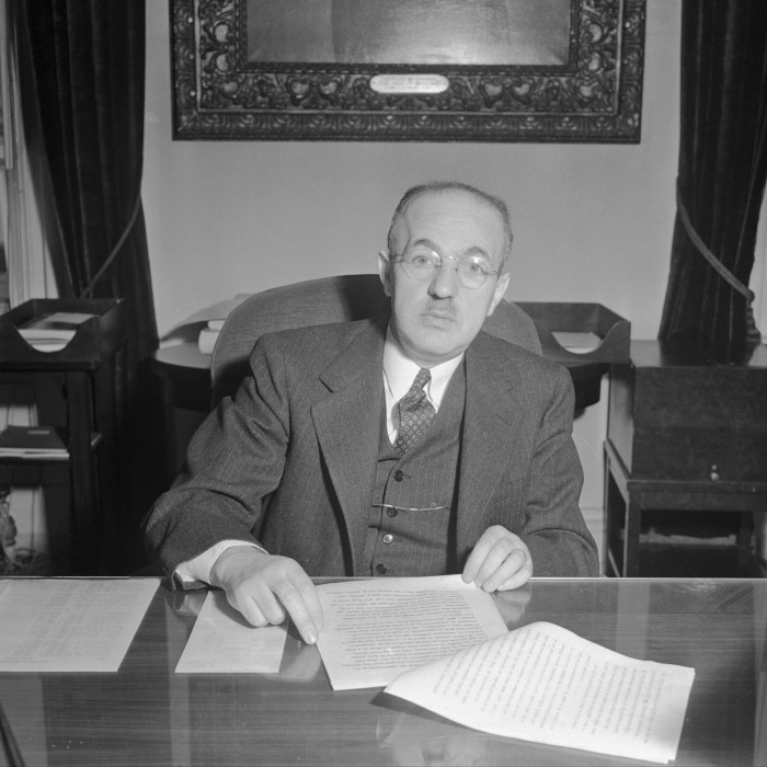 A man in a three-piece suit and wire rim spectacles sits at a desk, with a document in front on him