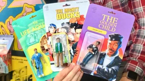 Wicked Joyful A man holding three home-made action figures. They look like real, professional packaged toys. The figures are all from movies - Eddie Murphy in The Golden Child, Bill Murray in The Life Aquatic, and Emilio Estevez in The Mighty Ducks.
