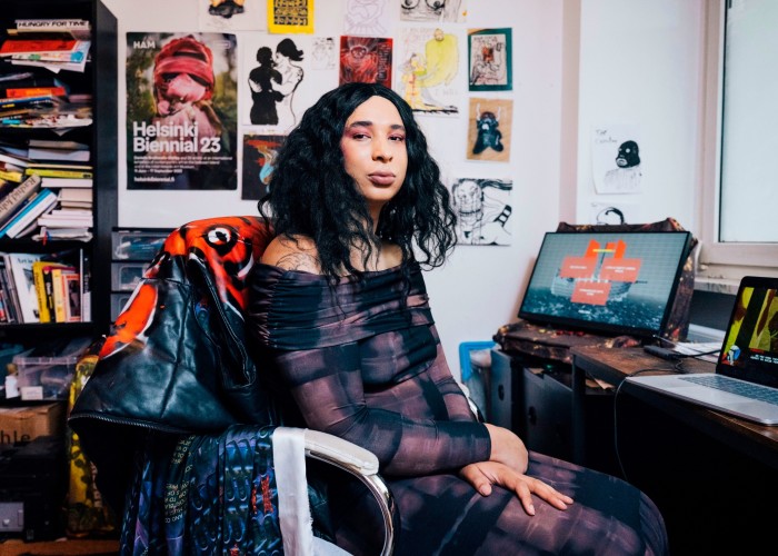 A trans woman sits at a desk in front of two computer screens, with artworks, posters and sketches pinned to a white wall behind her, along with a black shelving unit filled with books