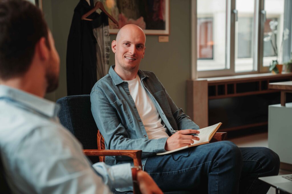Two colleagues are engaged in a conversation in a bright and modern office space. One person, holding a notebook and pen, is smiling while listening attentively. The setting is relaxed and professional, with artwork and natural light in the background.