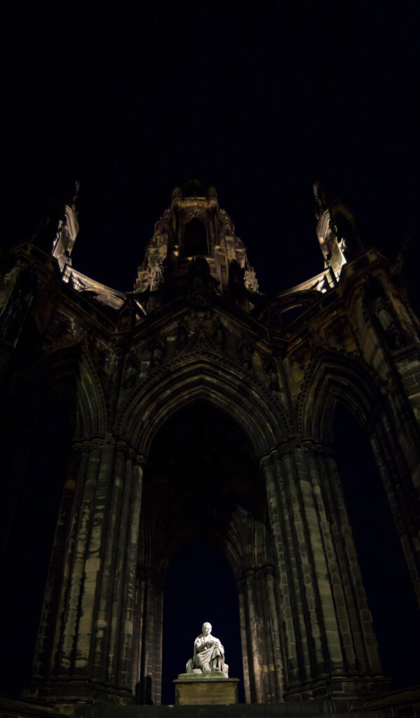 A photo looking up at the Scott Monument at night, with Scott illuminated at the bottom of the frame.