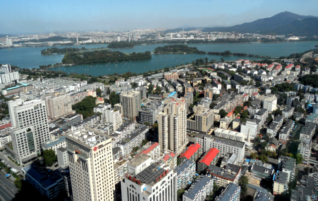 A photo of Nanjing city from above. In the foreground are the tops of skyscrapers and the rooves of buildings. In the background are a lake and a mountain.