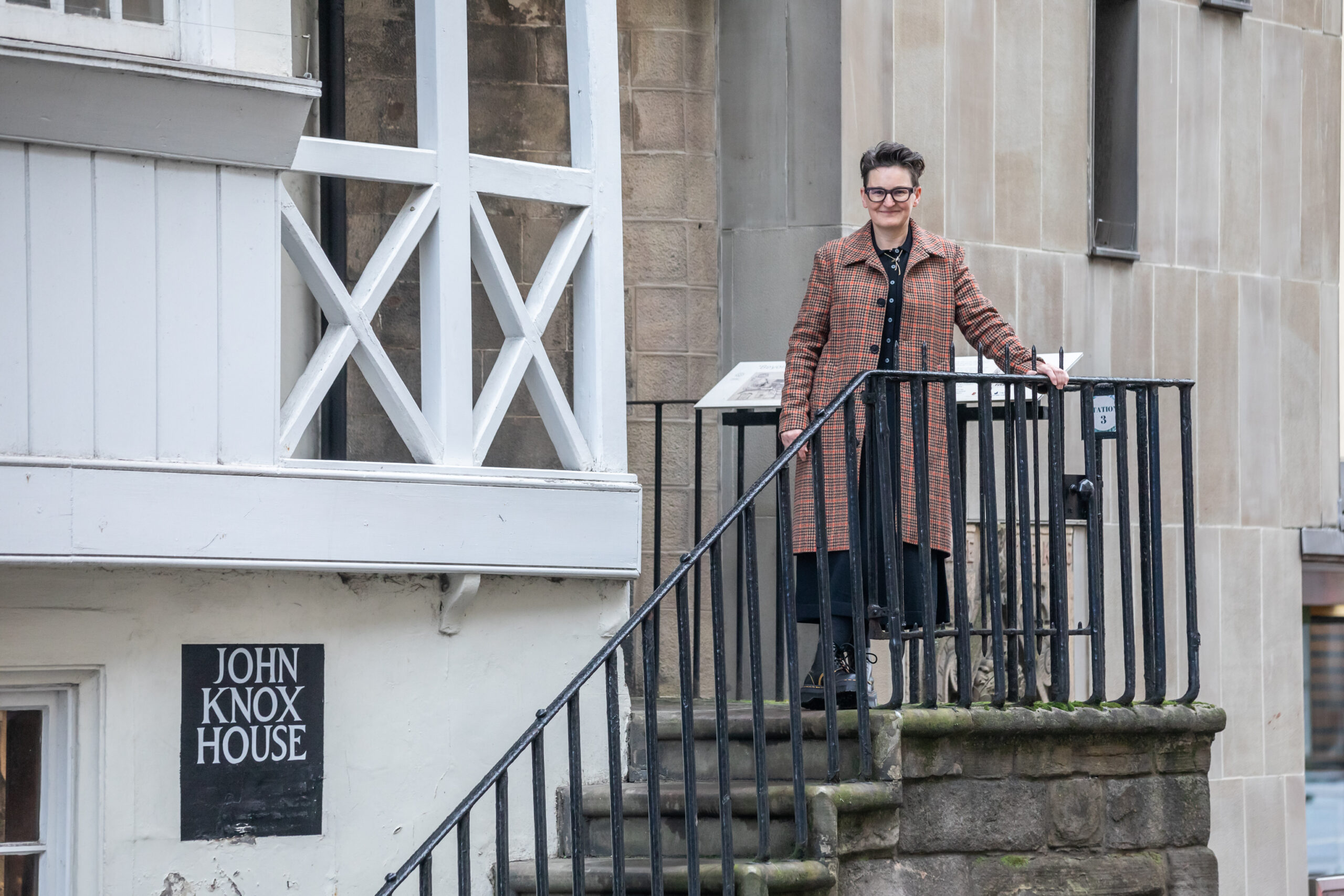 Mary Paulson-Ellis, a white woman with dark hair wearing a black outfit and an orange checked coat stands on the outside stone steps of a medieval building made of white wood.