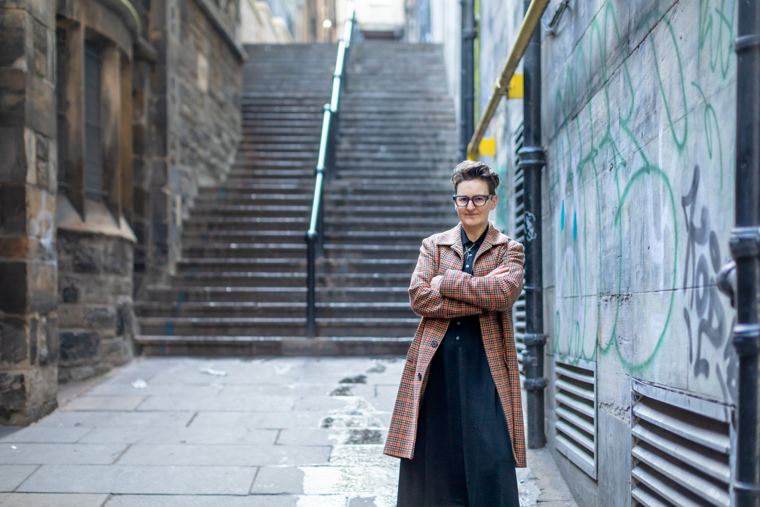 Mary Paulson-Ellis, a white woman with dark hair wearing a black outfit and an orange checked coat stands in a graffitied Edinburgh close with stone steps rising behind her.