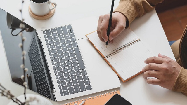 person writing notes in note book on desk with laptop