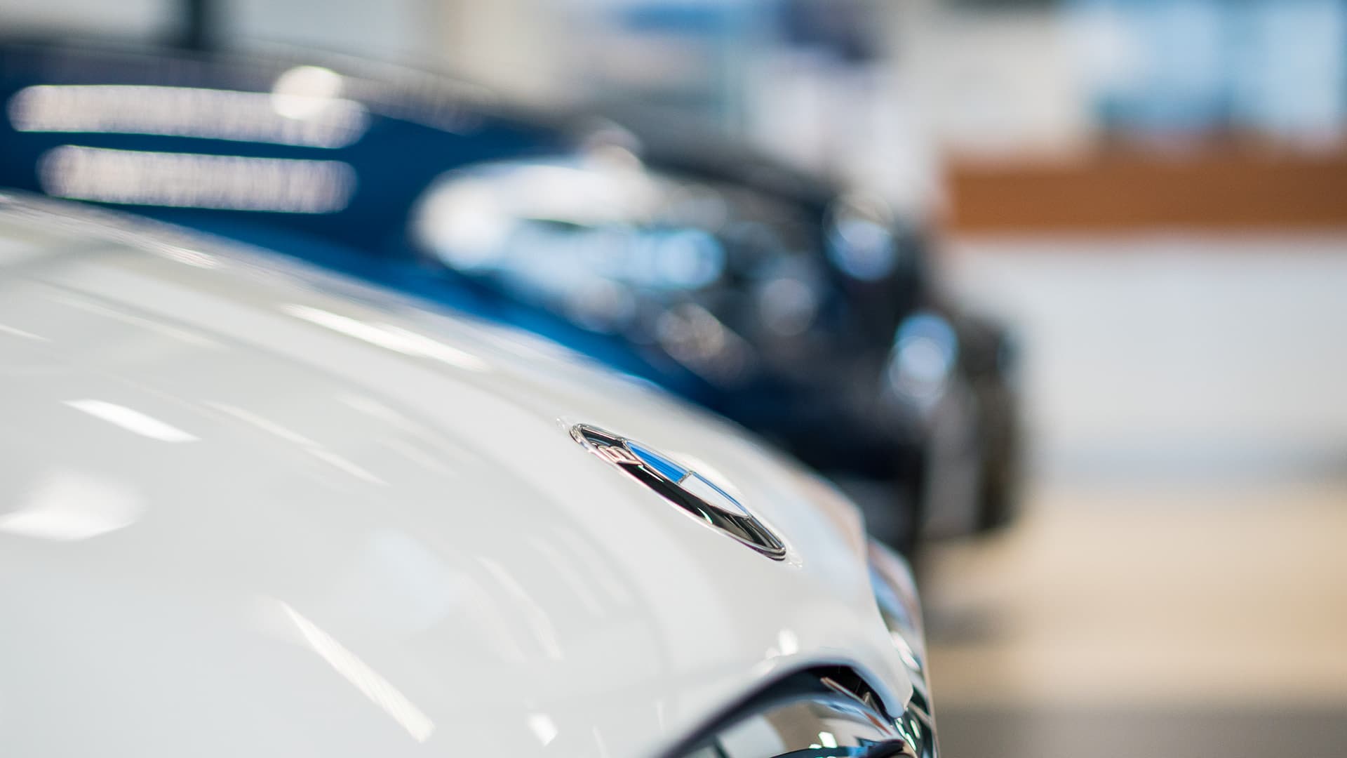 Nærbillede af en hvid bils motorhjelm med et sløret BMW-logo ved siden af andre biler, der er ude af fokus, i et lyst showroom, hvilket skaber en elegant, moderne stemning.