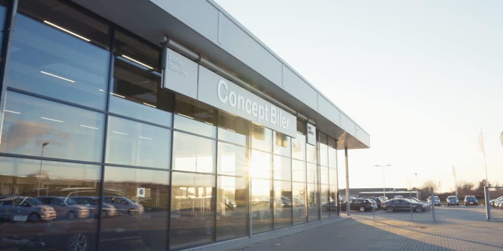 En moderne bilforhandler med store glasvinduer, der reflekterer sollyset, med skiltet "Concept Biler". Der er parkerede biler på parkeringspladsen.