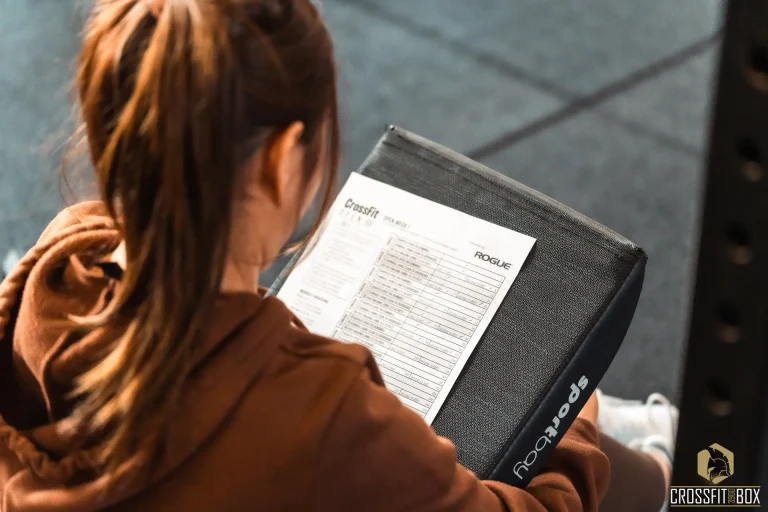 Een vrouwelijk lid zit op de grond met een scorekaart op haar schoot, terwijl ze de jurering doet voor een CrossFit-kwalificatiewedstrijd.