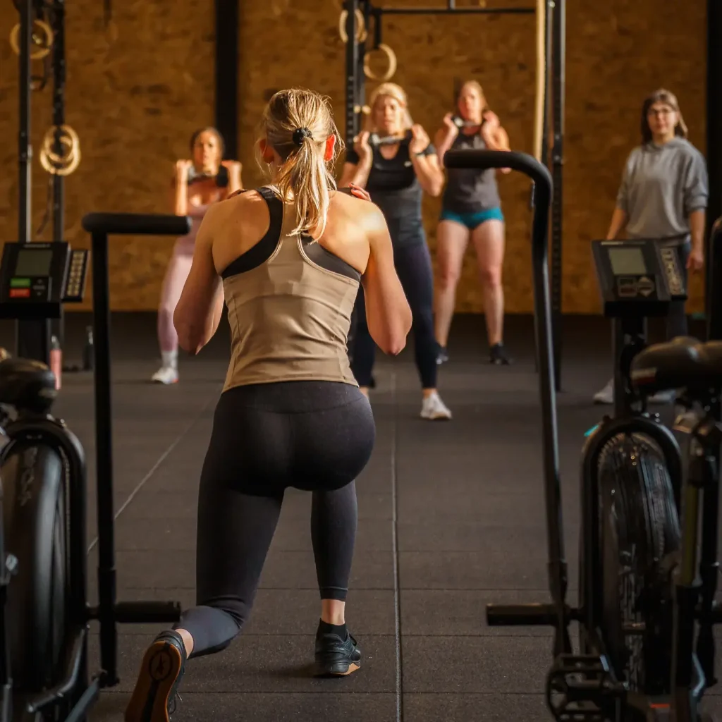 Een groep sporters volgt samen een oefening met een dumbbell in een groepsles, terwijl de coach aan de rechterkant de les begeleidt.