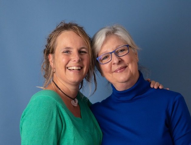 Simone en Inge, twee vrouwen in kleurrijke truien, lachen samen voor een foto met de hoofden tegen elkaar.