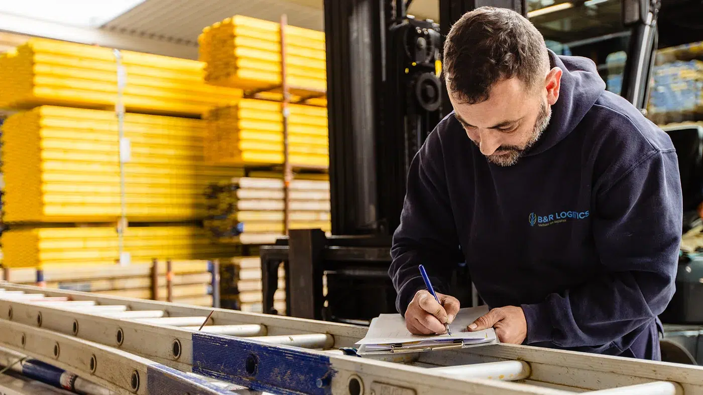 Een magazijnmedewerker van B&R Bouwgroep vult een piklijst in bij een stapel gele bekistingsbalken