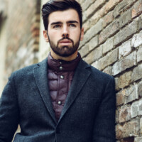 Young bearded man, model of fashion, standing in urban background wearing british elegant suit. Guy with beard and modern hairstyle in the street.