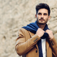Young man wearing winter clothes in the street. Young bearded guy with modern hairstyle with coat, scarf, blue jeans and t-shirt.
