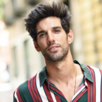 Attractive young man with dark hair and modern hairstyle wearing casual clothes in urban background.